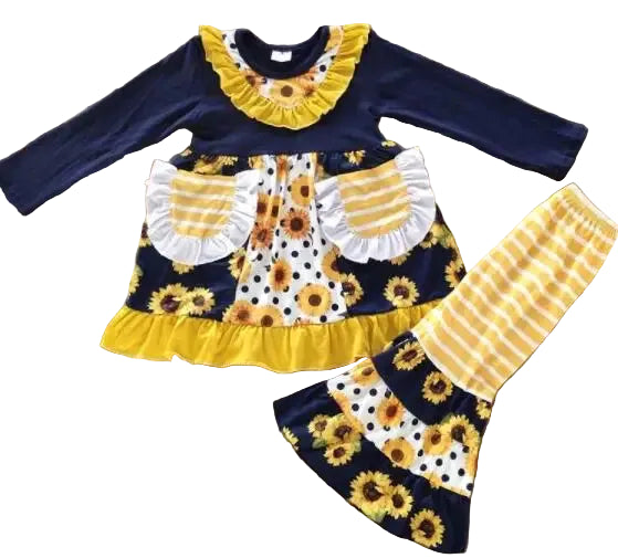 A toddler girl's two-piece clothing set laid flat on a wooden floor. The set includes a long-sleeve top and matching pants in navy blue, yellow, white, and black sunflower and polka dot patterns, with striped and ruffled details.