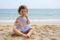 Toddler girl in a blue seersucker dress with pink embroidered bows, holding a water bottle by a window.