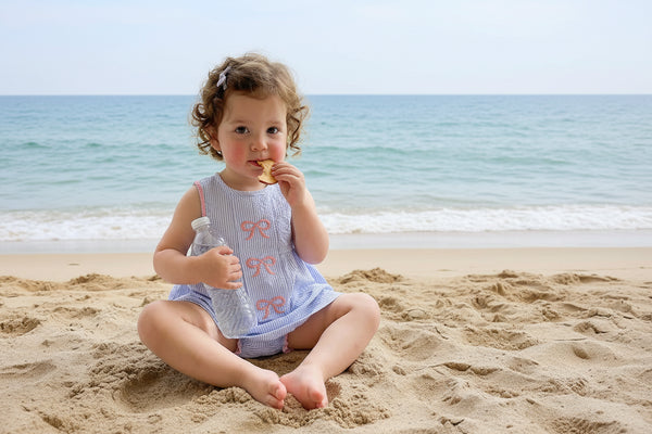 Toddler girl in a blue seersucker dress with pink embroidered bows, holding a water bottle by a window.