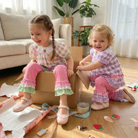 Two little girls in frilly boho plaid embroidered two-piece sets (pink and purple), playing with torn wrapping paper.