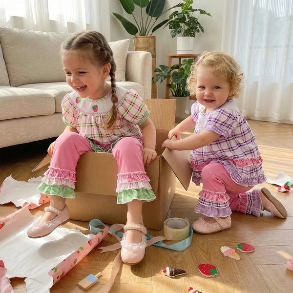 Two little girls in frilly boho plaid embroidered two-piece sets (pink and purple), playing with torn wrapping paper.