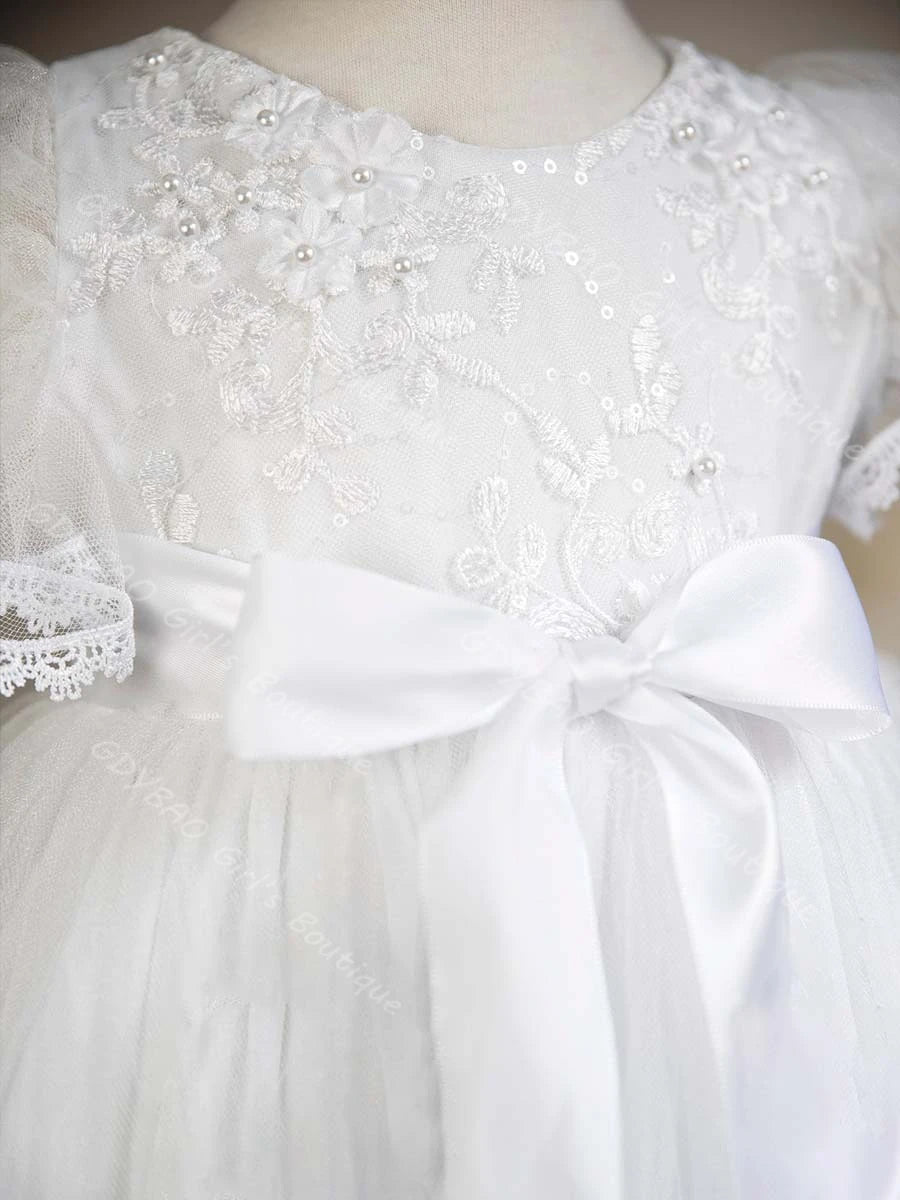 White baby christening gown featuring lace floral appliques, pearl beading, and a large satin bow at the waist.