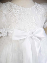 White baby christening gown featuring lace floral appliques, pearl beading, and a large satin bow at the waist.