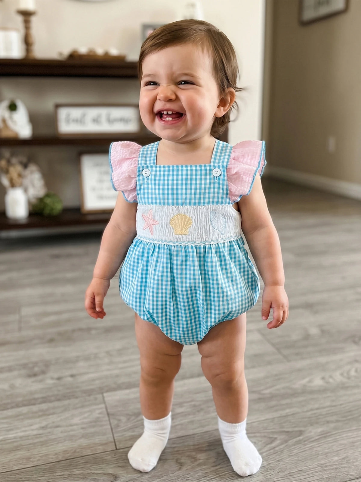 Smiling baby in blue gingham romper with pink flutter sleeves and sea creature embroidery.