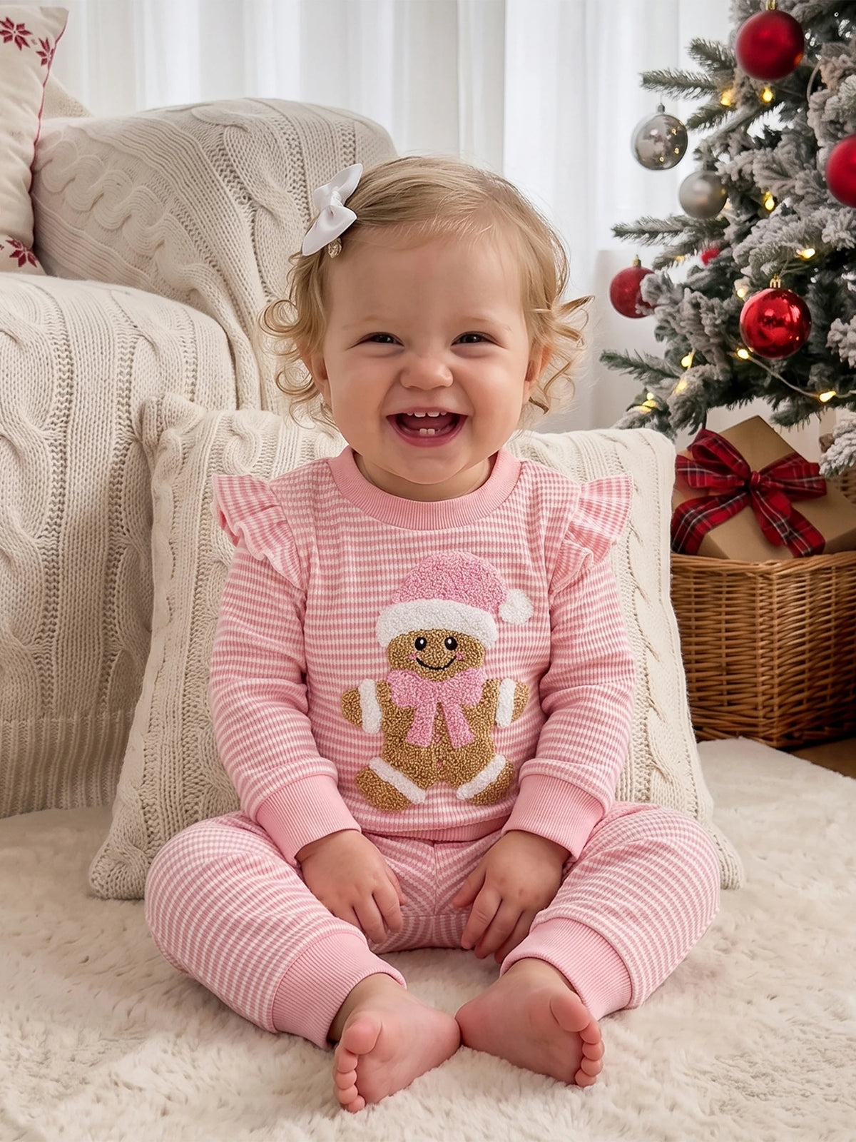 Toddler girl wearing pink and white striped waffle knit 2-piece Christmas set with Gingerbread Baby embroidery near decorated tree.