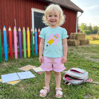 Toddler girl in light blue embroidered top with pencil bow and puff sleeves, pink striped shorts outdoors by a fence.