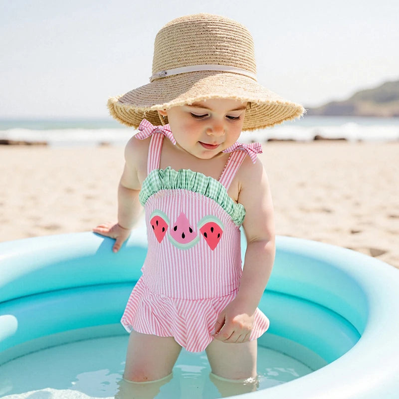 Adorable child in the 'Watermelon Baby' Cute One Piece Swimsuit playing in a kiddie pool at the beach, wearing a sun hat.