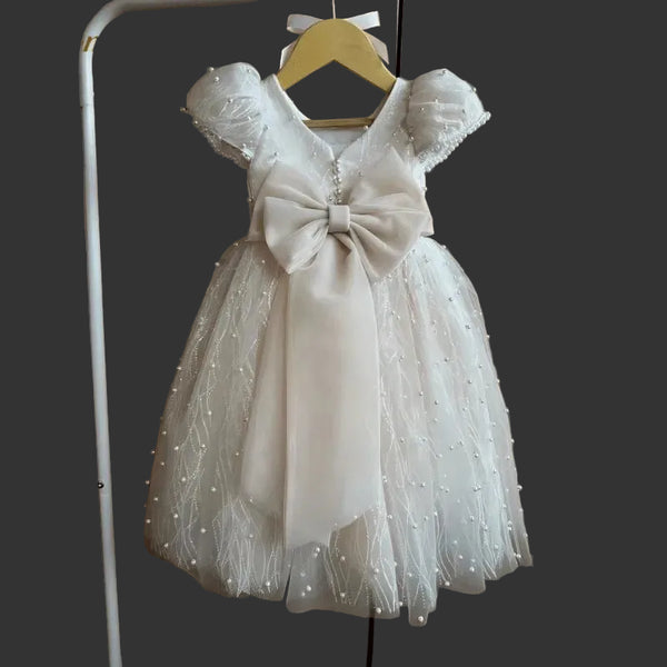 White children's dress with a large bow on a hanger against a neutral background