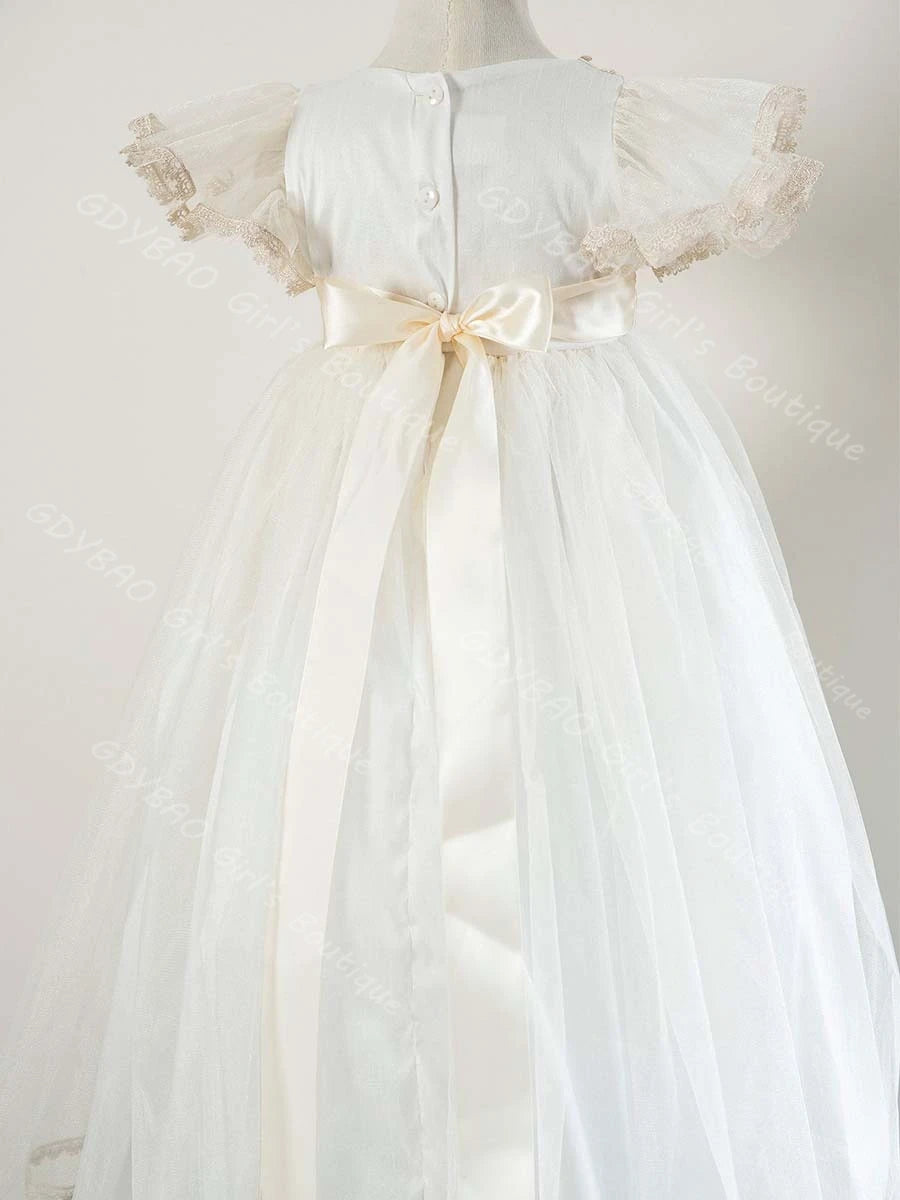 Back view of white baby girl christening gown featuring lace cap sleeves, satin ribbon sash bow, and floor-length tulle skirt.