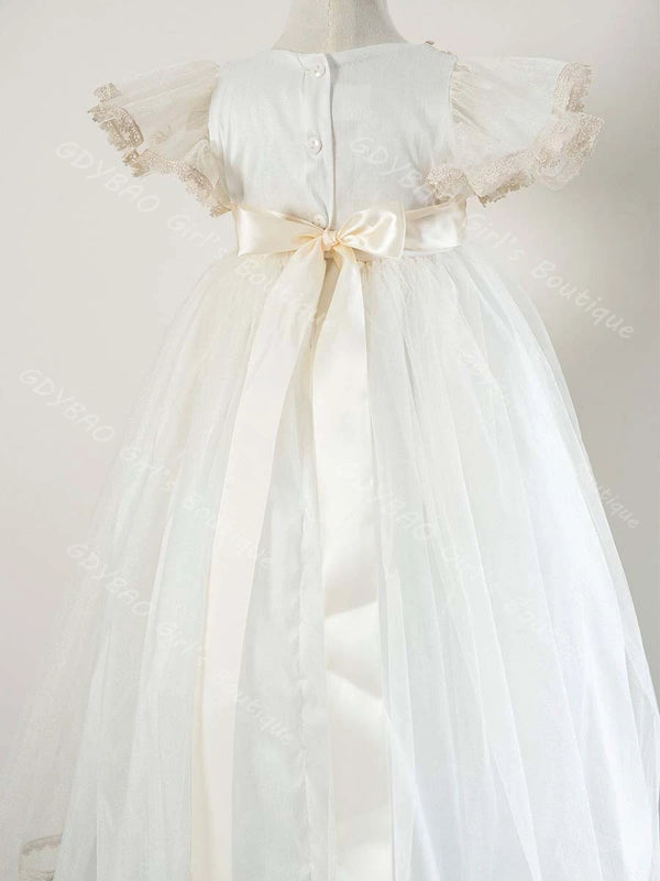 Back view of white baby girl christening gown featuring lace cap sleeves, satin ribbon sash bow, and floor-length tulle skirt.