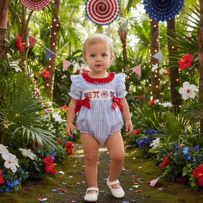 Baby in a patriotic outfit standing on a wooden floor with decorative flags in the background.