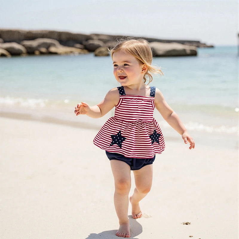 A happy young girl wearing the 'Stars and Stripes' 2 PC Dress and Bloomer Set, playing on the beach with a joyful expression, showcasing the red and white striped dress with navy blue accents.
