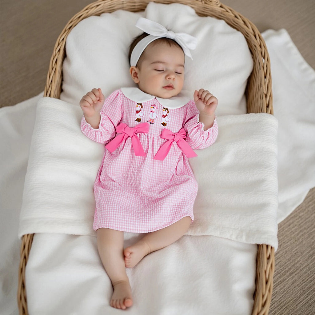 Baby wearing the 'Cowboy Goosies' Pink Checked Smocked Little Girls Dress, lying peacefully in a basket with a headband.