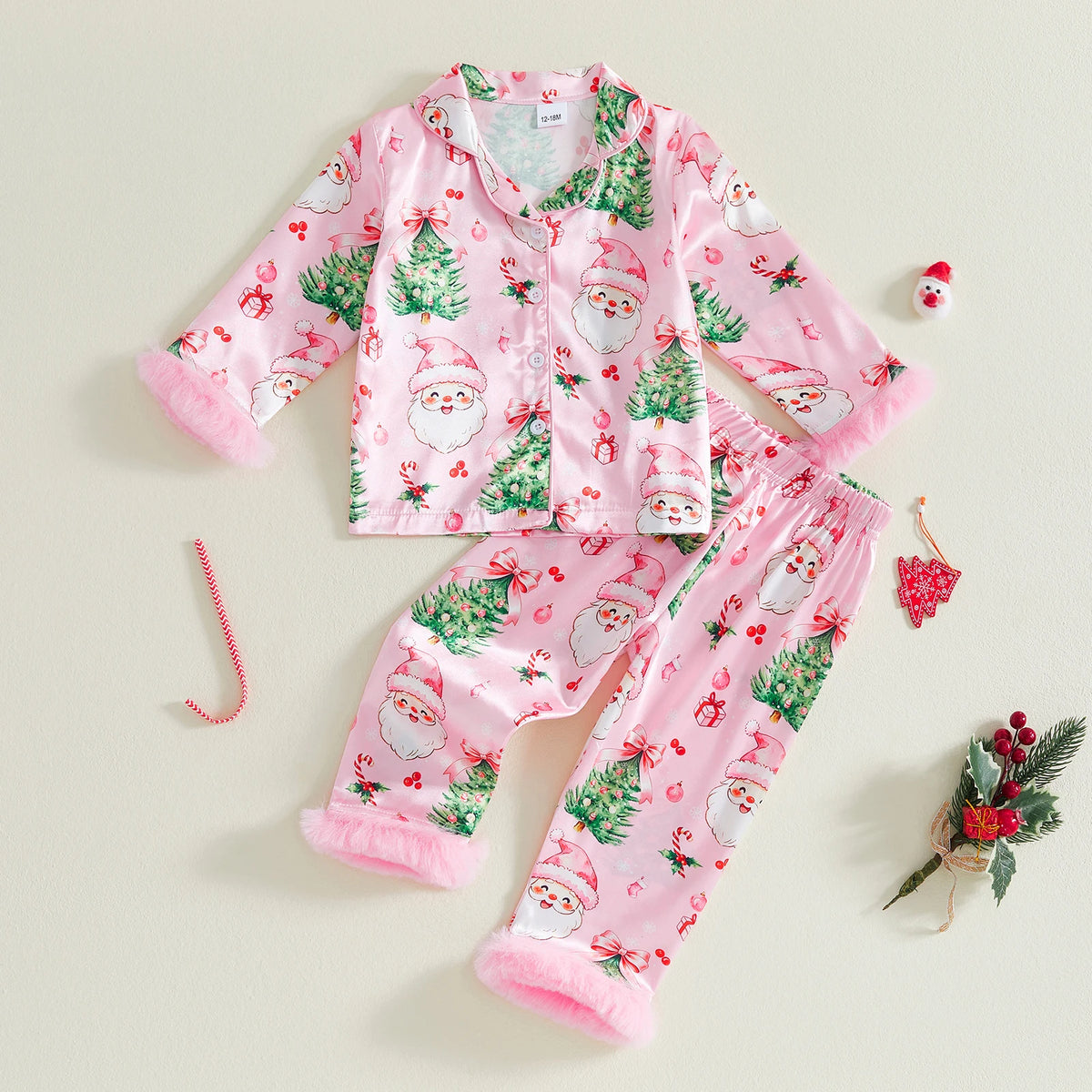Girls pink satin Christmas pajama set featuring Santa, trees, and bows with fuzzy cuffs. 2-piece button front lounge wear for holidays.