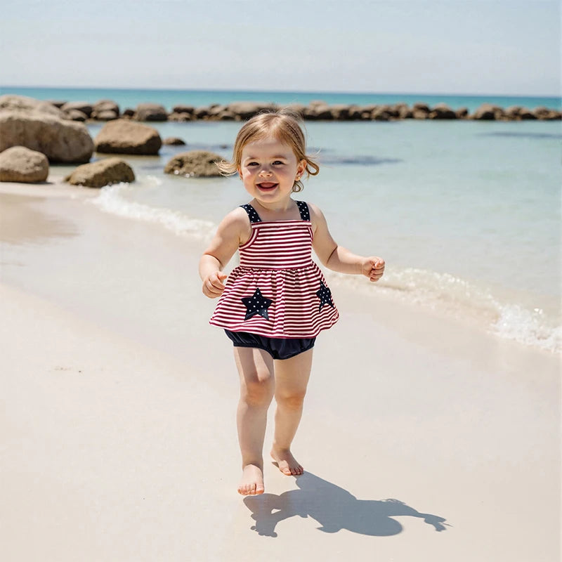 A cheerful little girl running on the beach, wearing the 'Stars and Stripes' 2 PC Dress and Bloomer Set, capturing the spirit of fun and adventure in warm summer weather.