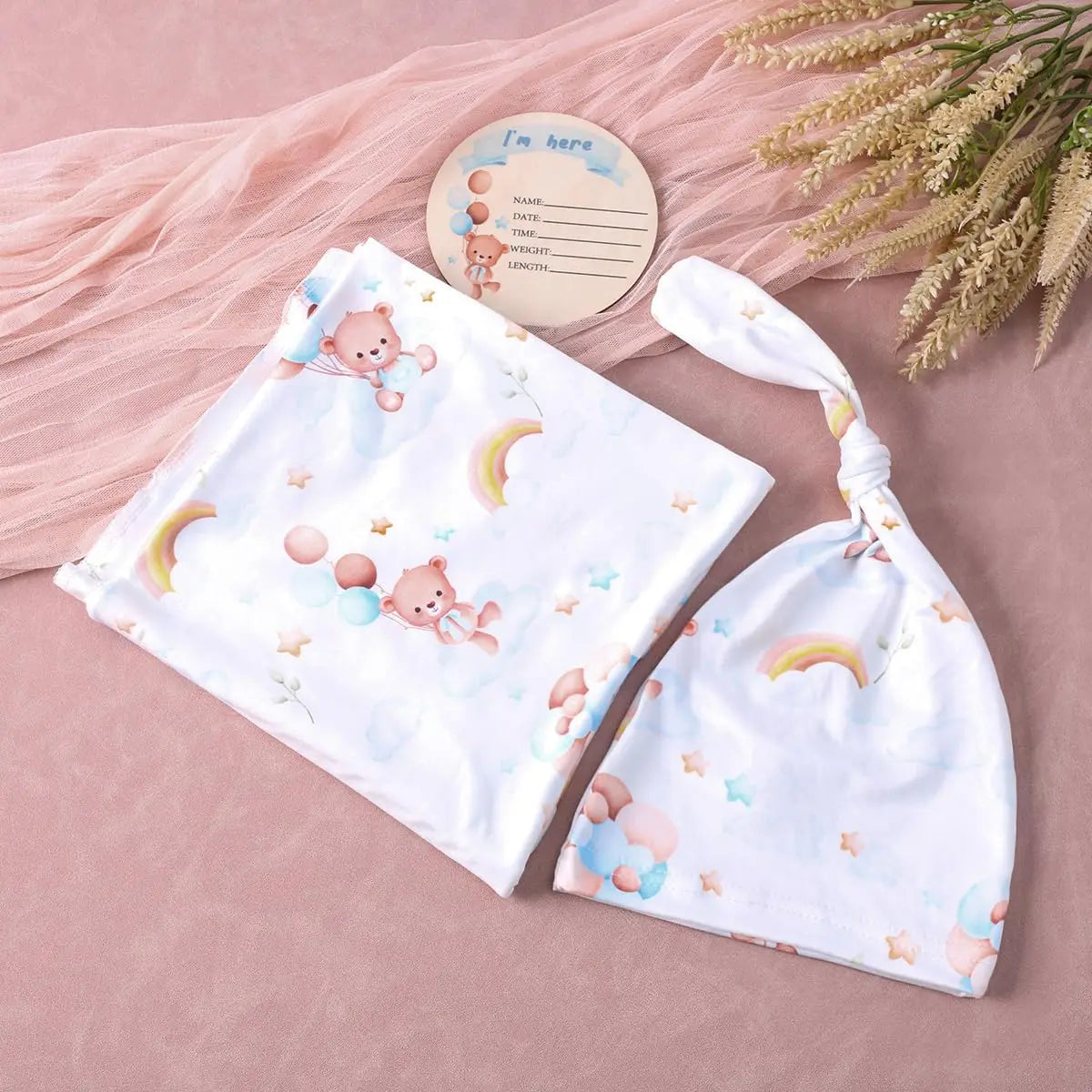 White baby swaddle and tied headband set featuring teddy bears, rainbows, and stars on a pink background with an "I'm here" tag.