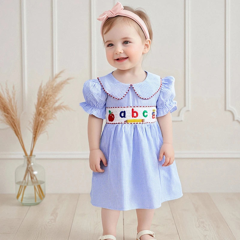 A toddler wearing the ABCs Embroidered Girls Dress - Peter Pan Collar, featuring playful embroidery and a Peter Pan collar, smiling in a charming indoor setting.