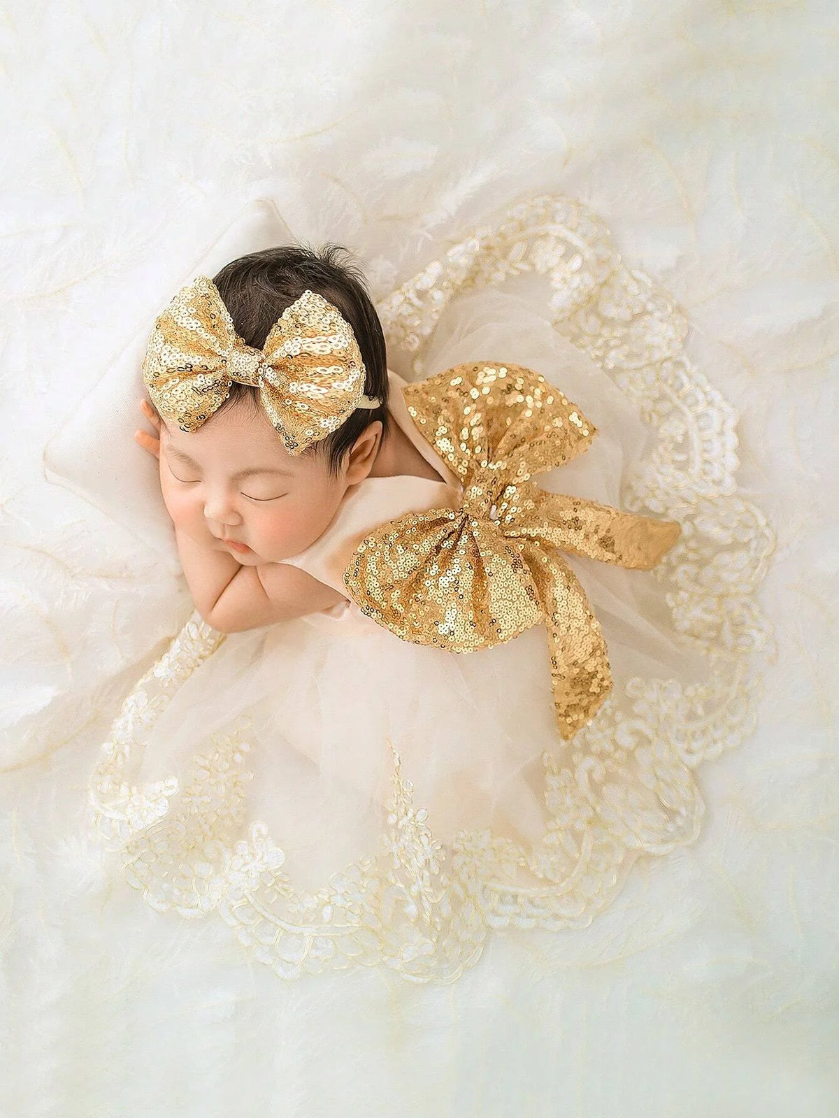 Sleeping infant wearing a gold sequined headband bow and an oversized sequined bow on a lace gown.