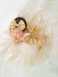 Sleeping infant wearing a gold sequined headband bow and an oversized sequined bow on a lace gown.