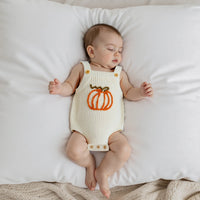 Sleeping baby wearing cream knit romper overalls with an orange embroidered pumpkin on the chest. Sleeveless seasonal baby outfit.