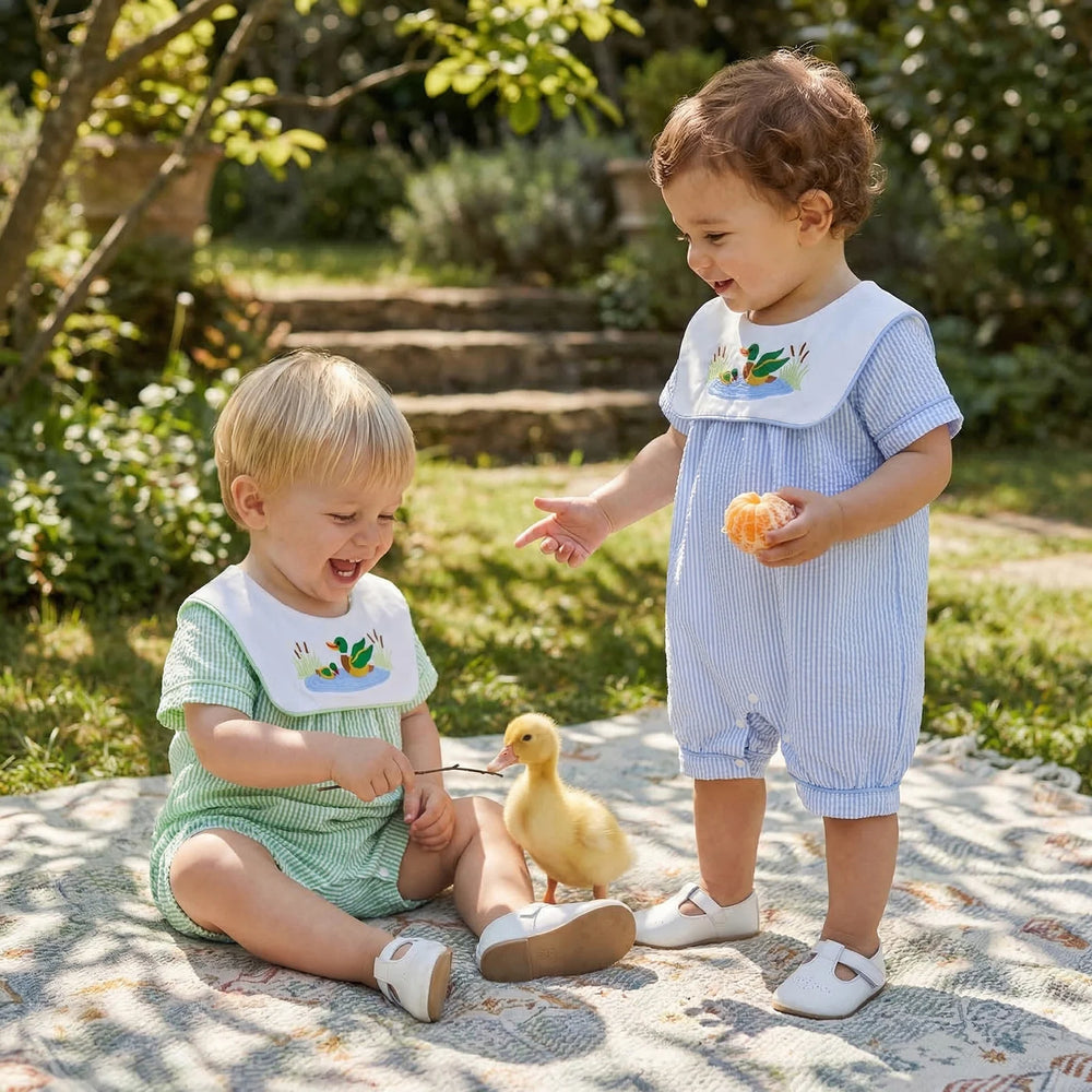 Two babies wear Little Ducky bibbed seersucker rompers in Boca Blue and Green, with embroidered mallard ducks.