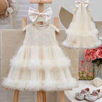 A sleeveless cream tulle dress for toddlers or girls, featuring a sequined bodice, layered ruffle skirt, and a large matching bow headband. The dress is displayed on a wooden hanger stand, with a smaller inset view of the dress on the upper right and a decorative floral corner on the lower right.
