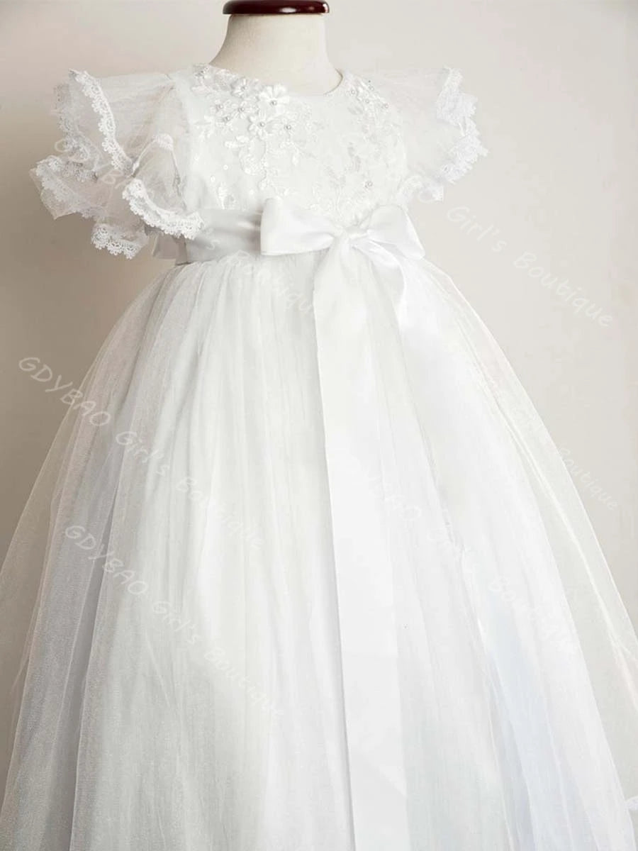 White baby girl christening gown with lace ruffle sleeves, floral beading, and satin bow sash on floor-length tulle skirt.