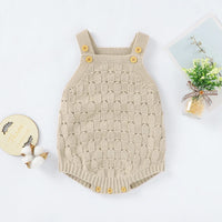 A baby knit romper in a light beige color with a textured pattern, featuring yellow buttons on the shoulder straps and crotch. The romper is displayed in a flat-lay arrangement on a white background, styled with decorative props including cotton bolls, a small wooden sign, and a miniature potted plant.