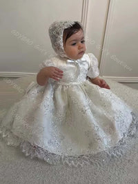 Baby girl wearing white sequined lace baptism dress with matching bonnet, ruffles, and bow for formal occasions.
