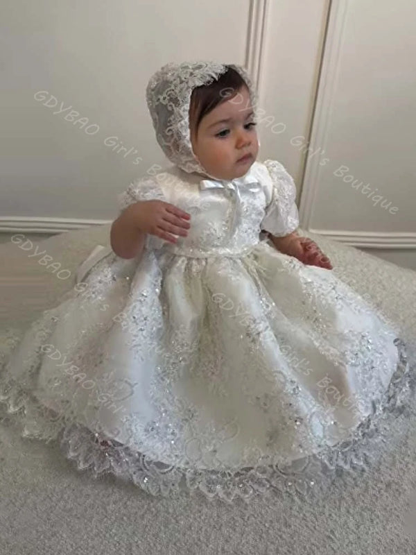 Baby girl wearing white sequined lace baptism dress with matching bonnet, ruffles, and bow for formal occasions.