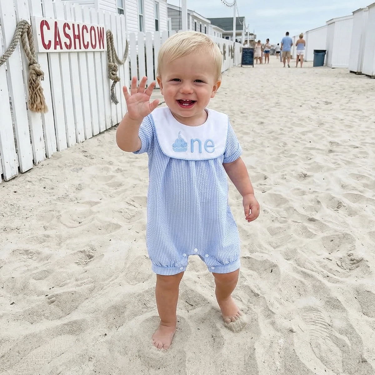 Child in a blue outfit with 'one' on it, standing on a sandy beach.