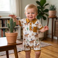 Baby boy in polo-style romper with western/fishing print, tan collar and pocket, indoors beside a cactus.