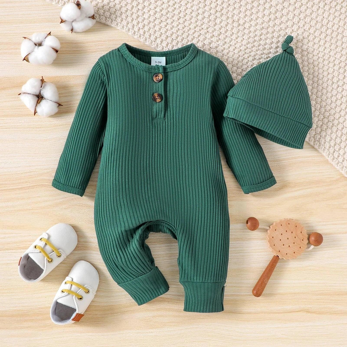 green ribbed romper and matching hat for baby laid flat with white baby shoes and wooden rattle