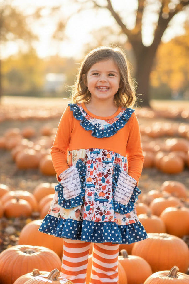 A toddler girl's long-sleeve dress and matching striped leggings set laid flat on a dark wood floor. The dress has a solid orange long sleeve and yoke, a blue polka-dot ruffled collar, and a tiered skirt with a white background and a colorful autumn leaf and pumpkin floral pattern, featuring two front pockets.