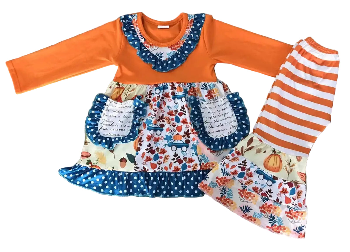 A toddler girl's long-sleeve dress and matching striped leggings set laid flat on a dark wood floor. The dress has a solid orange long sleeve and yoke, a blue polka-dot ruffled collar, and a tiered skirt with a white background and a colorful autumn leaf and pumpkin floral pattern, featuring two front pockets.