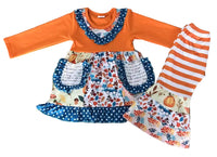 A toddler girl's long-sleeve dress and matching bell-bottom pants set laid flat on a white background. The dress has an orange long-sleeve top, a blue polka-dot ruffled collar, and a floral apron-style skirt with pockets.