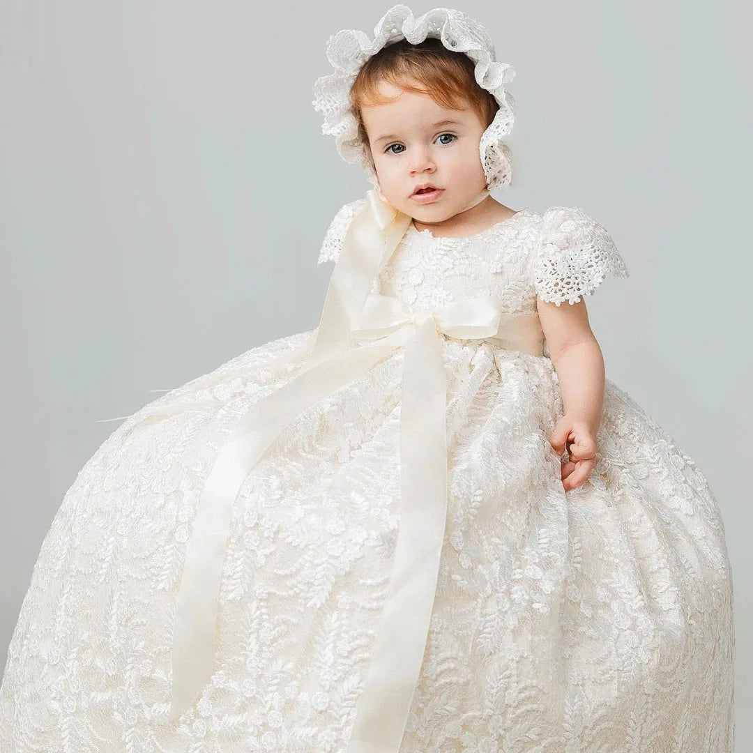 Baby wearing ivory lace full-length gown with satin bow and matching bonnet for baptism, weddings, or portraits.