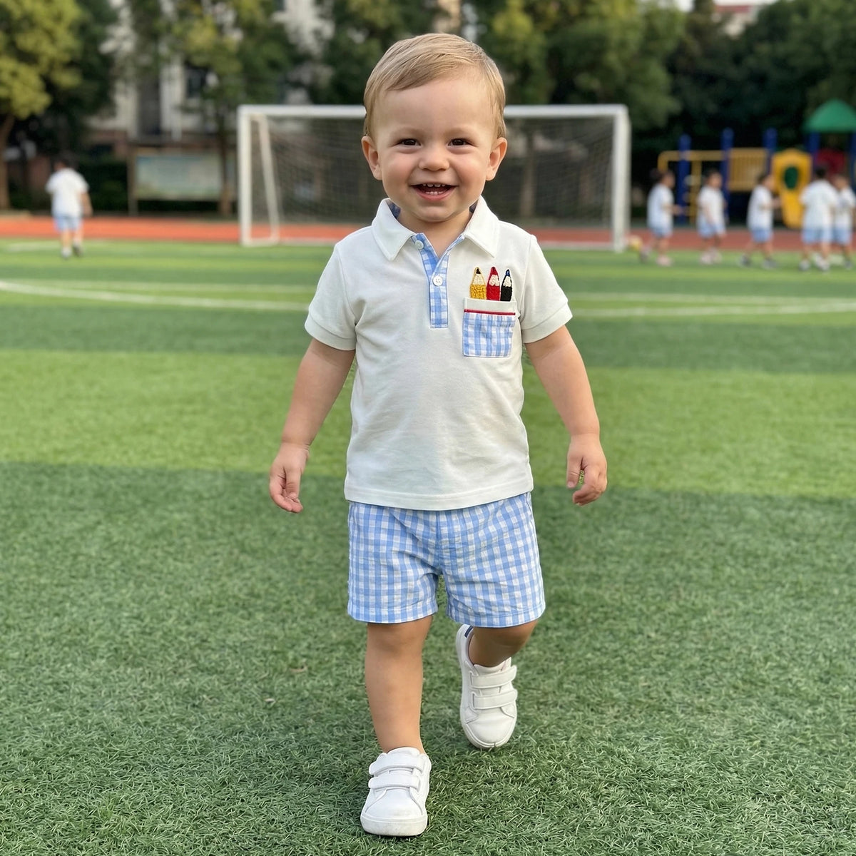 Toddler boy in white polo with pencil embroidery and blue gingham shorts on a grassy field.
