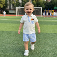 Toddler boy in white polo with pencil embroidery and blue gingham shorts on a grassy field.