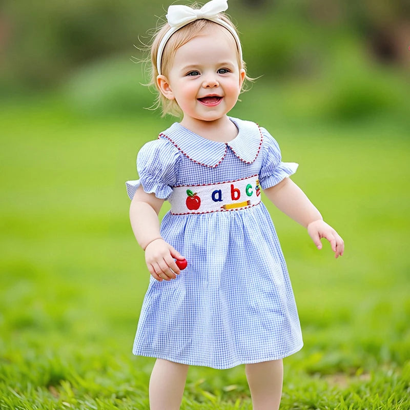 A happy little girl outdoors in the ABCs Embroidered Girls Dress - Peter Pan Collar, showcasing colorful embroidery and a soft blue seersucker design.