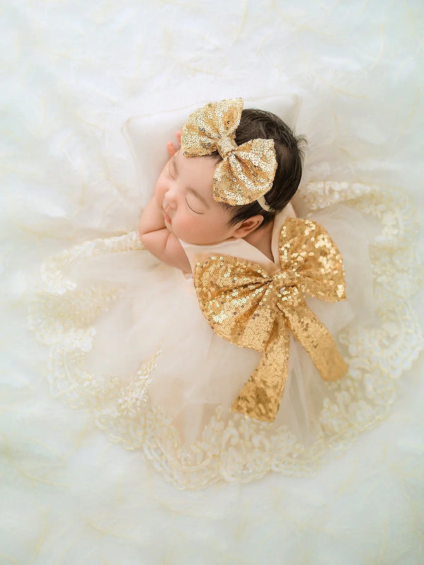 Infant girl in gold sequin oversized bow headband with matching bow dress and lace accents.