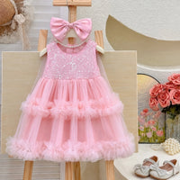 A pink sleeveless baby girl's party dress with a sequined bodice, a large matching pink bow, and a tiered tulle skirt, displayed on a wooden stand.