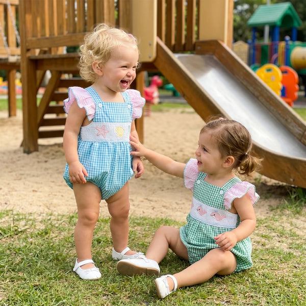 Two toddlers in seashore romper with embroidered sea creatures and pink flutter sleeves, at a playground.