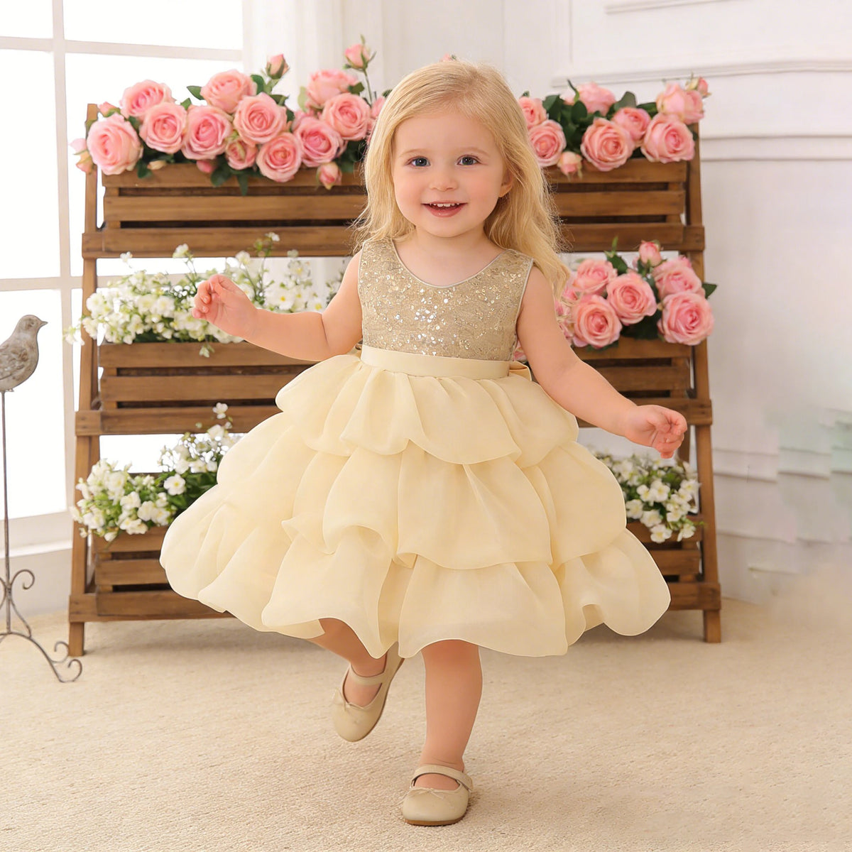 Toddler girl wearing a gold sequined bodice dress with a tiered cream skirt and detachable bow, set against pink roses.