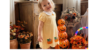 A young toddler girl stands in a decorated indoor room wearing a pale yellow short-sleeve dress with a white collar and small pumpkin appliques near the hem. She is posed in front of Halloween decorations including stacked jack-o'-lanterns, autumn flowers, a spiderweb decoration, and a glowing hoop light.