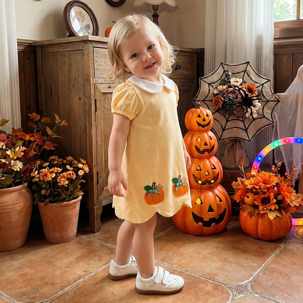 A young toddler girl stands in a decorated indoor room wearing a pale yellow short-sleeve dress with a white collar and small pumpkin appliques near the hem. She is posed in front of Halloween decorations including stacked jack-o'-lanterns, autumn flowers, a spiderweb decoration, and a glowing hoop light.