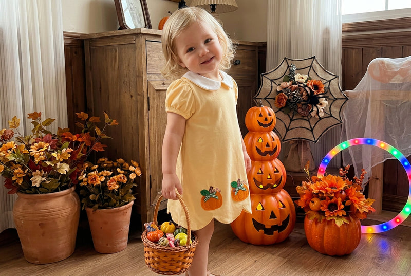 A young toddler girl stands in a decorated indoor room wearing a pale yellow short-sleeve dress with a white collar and small pumpkin appliques near the hem. She is posed in front of Halloween decorations including stacked jack-o'-lanterns, autumn flowers, a spiderweb decoration, and a glowing hoop light.