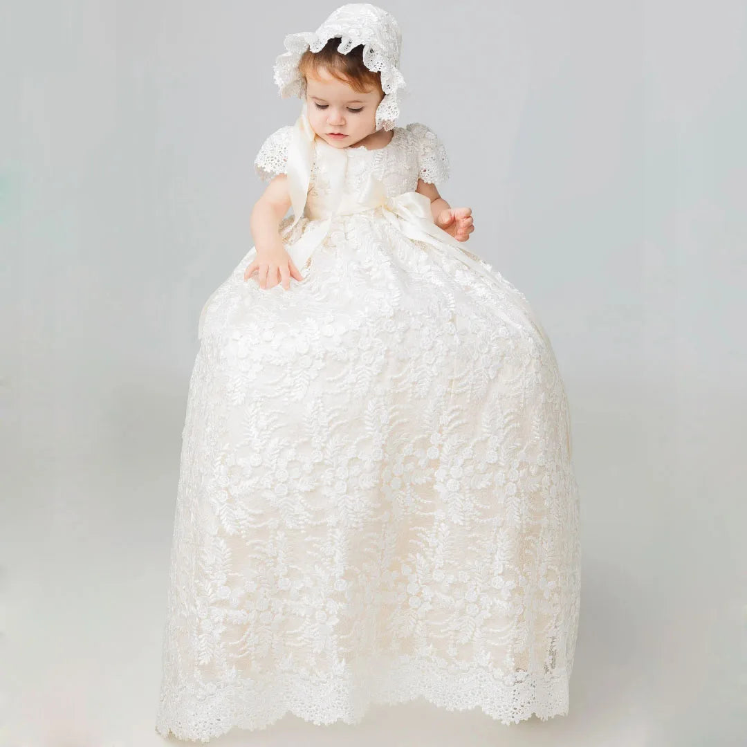 Baby wearing elegant ivory full-length lace gown with matching bonnet, satin ribbon bow, and floral appliques for baptism or wedding.