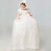 Baby wearing elegant ivory full-length lace gown with matching bonnet, satin ribbon bow, and floral appliques for baptism or wedding.