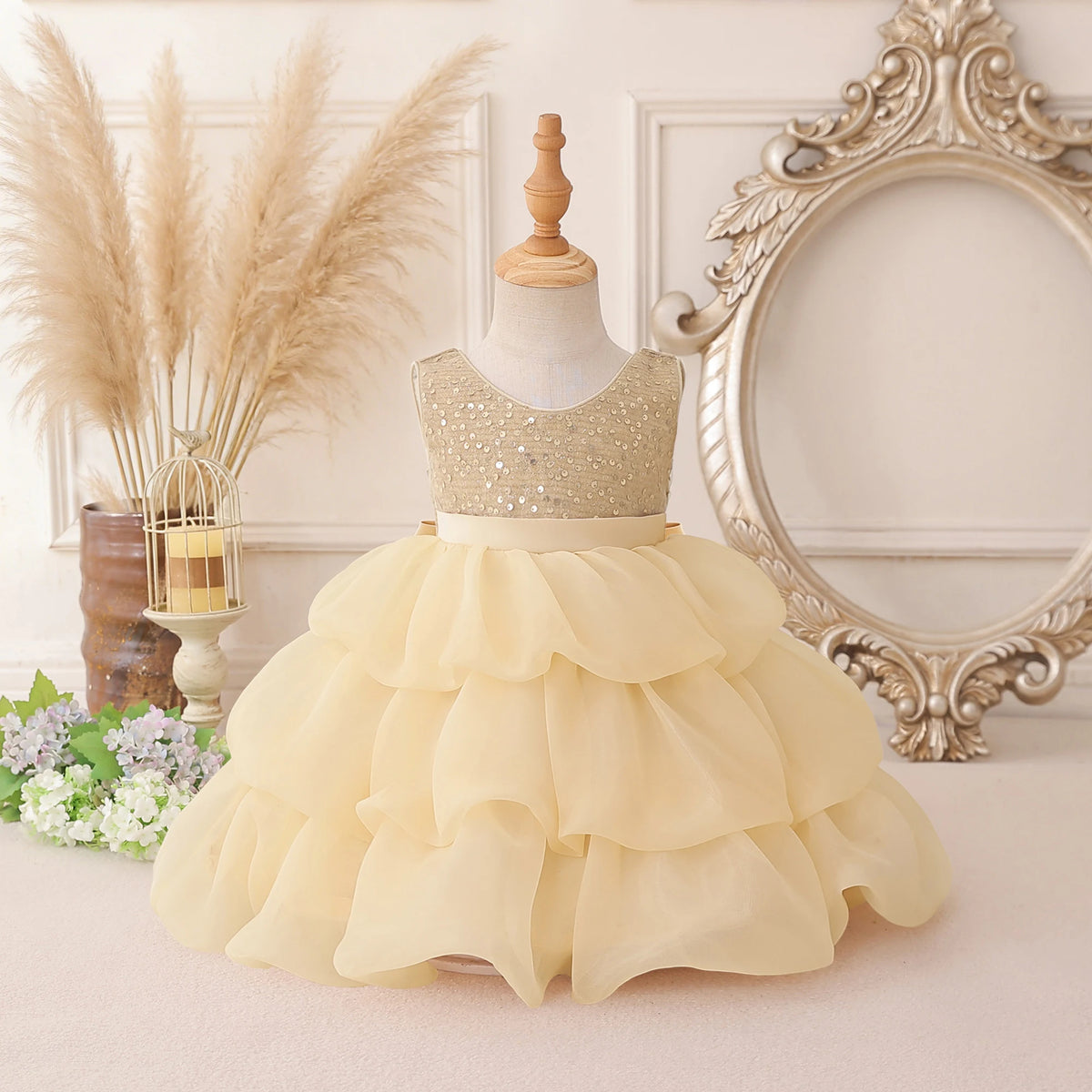 A toddler girl's formal dress displayed on a dress form mannequin, featuring a champagne gold sequined bodice and a voluminous cream tulle tiered skirt, set against a vintage-style room backdrop with a mirror and pampas grass.